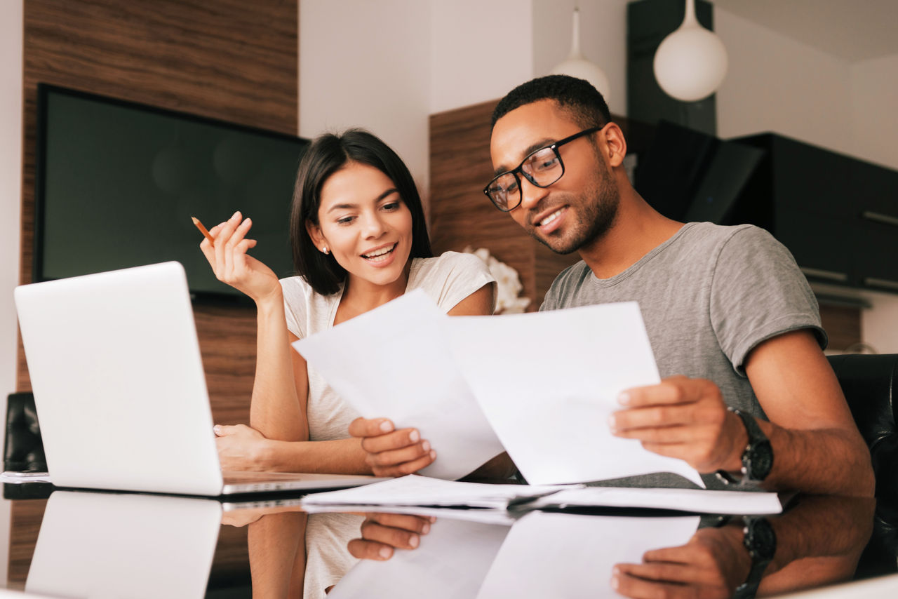 Photo of cheerful loving young couple using laptop and analyzing their finances with documents. Look at papers.